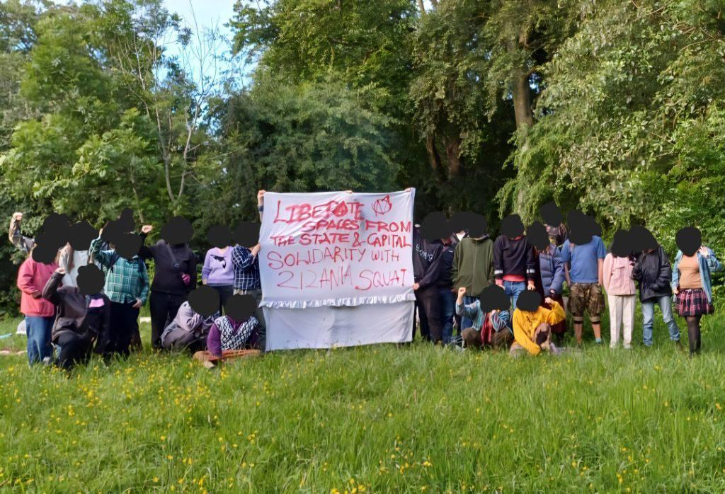 a group of 30 people holding a banner in a park holding a banner. Their faces are blacked out. The banner reads "LIBERATE SPACES FROM THE STATE AND CAPITAL, SOLIDARITY WITH ZIZANIA SQUAT"